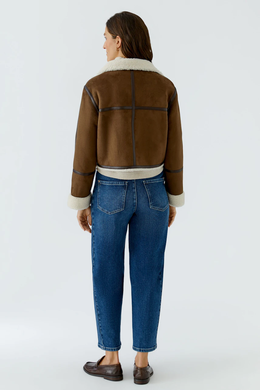 Lederimitatjacke: Frau in brauner Jacke mit hellem Kragen, kombiniert mit blauen Jeans und flachen Schuhen.