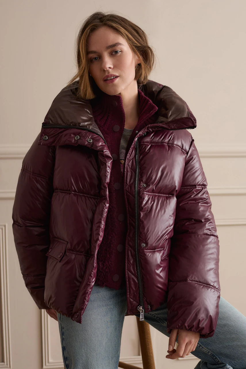 Frau in bordeauxfarbener Outdoorjacke, kombiniert mit Strickpullover und Jeans, steht lässig im Raum.