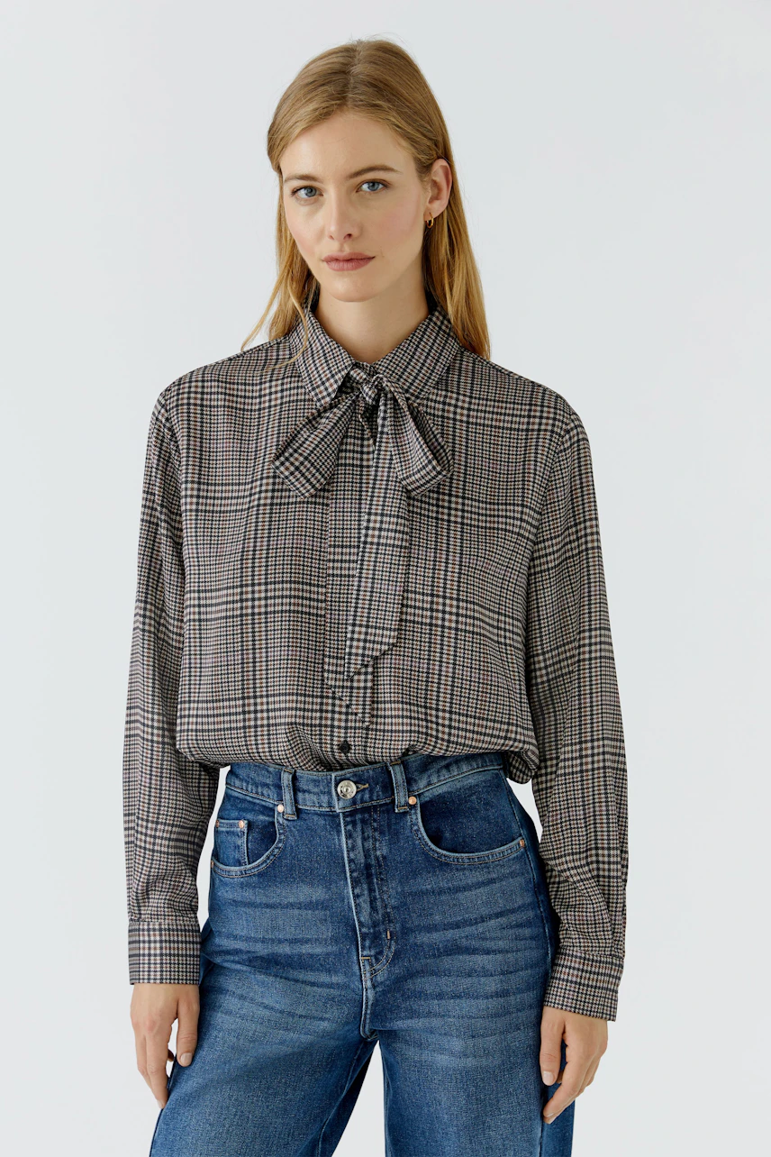 Woman in a checkered pussy-bow blouse and blue jeans, standing confidently.