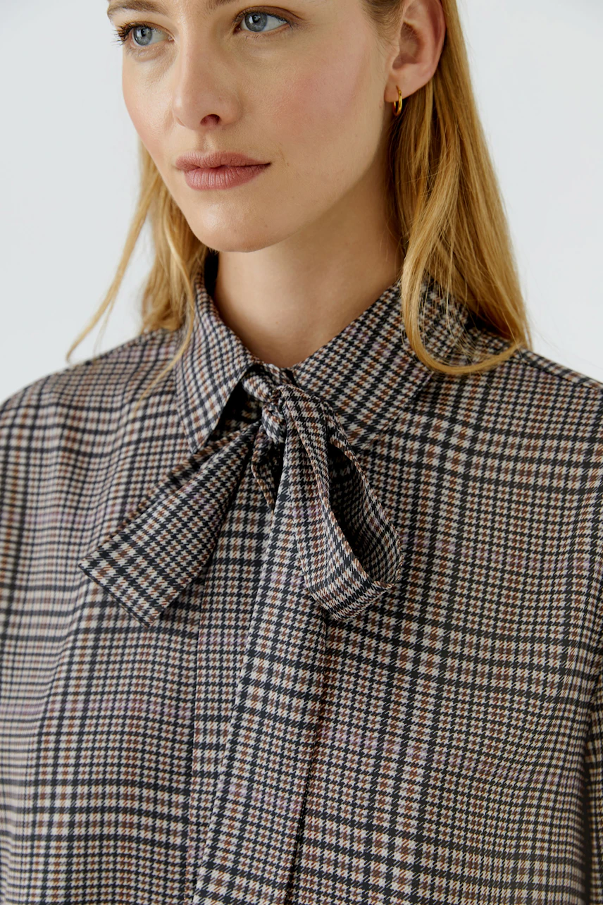 Woman in checkered pussy-bow blouse, looking poised. The blouse features a classic bow tie detail.