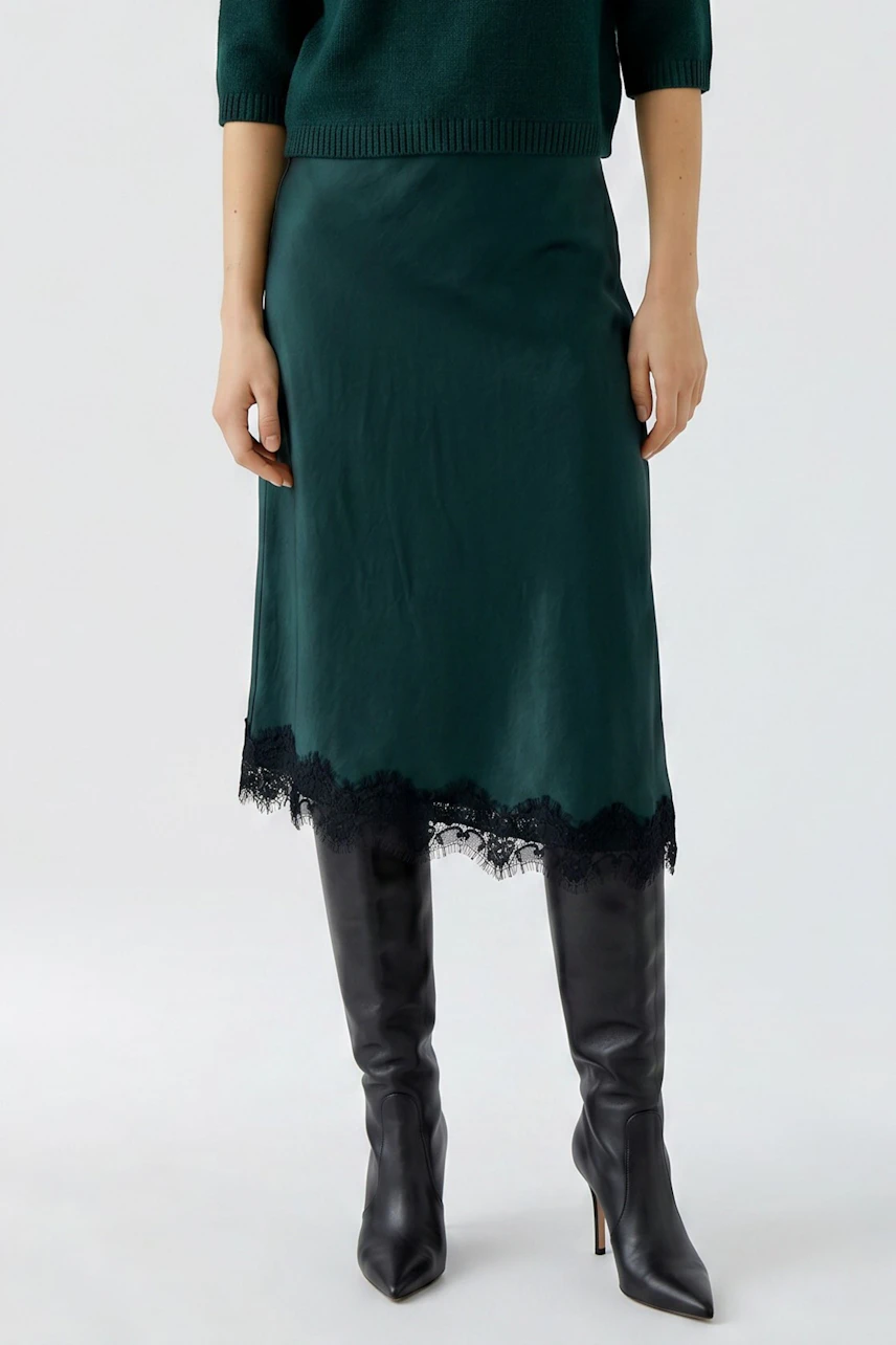 Woman in green pencil skirt with lace hem, paired with black boots, showcasing a chic and modern style.