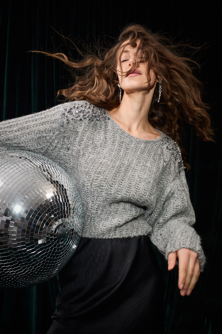 Woman in a gray pullover and black pencil skirt, holding a disco ball, with dynamic hair movement.