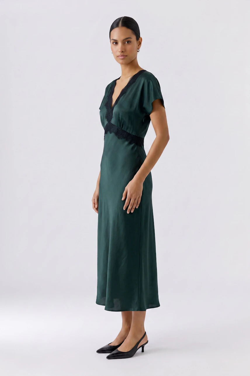 Woman in a green dress with black lace detail, standing confidently in black heels.