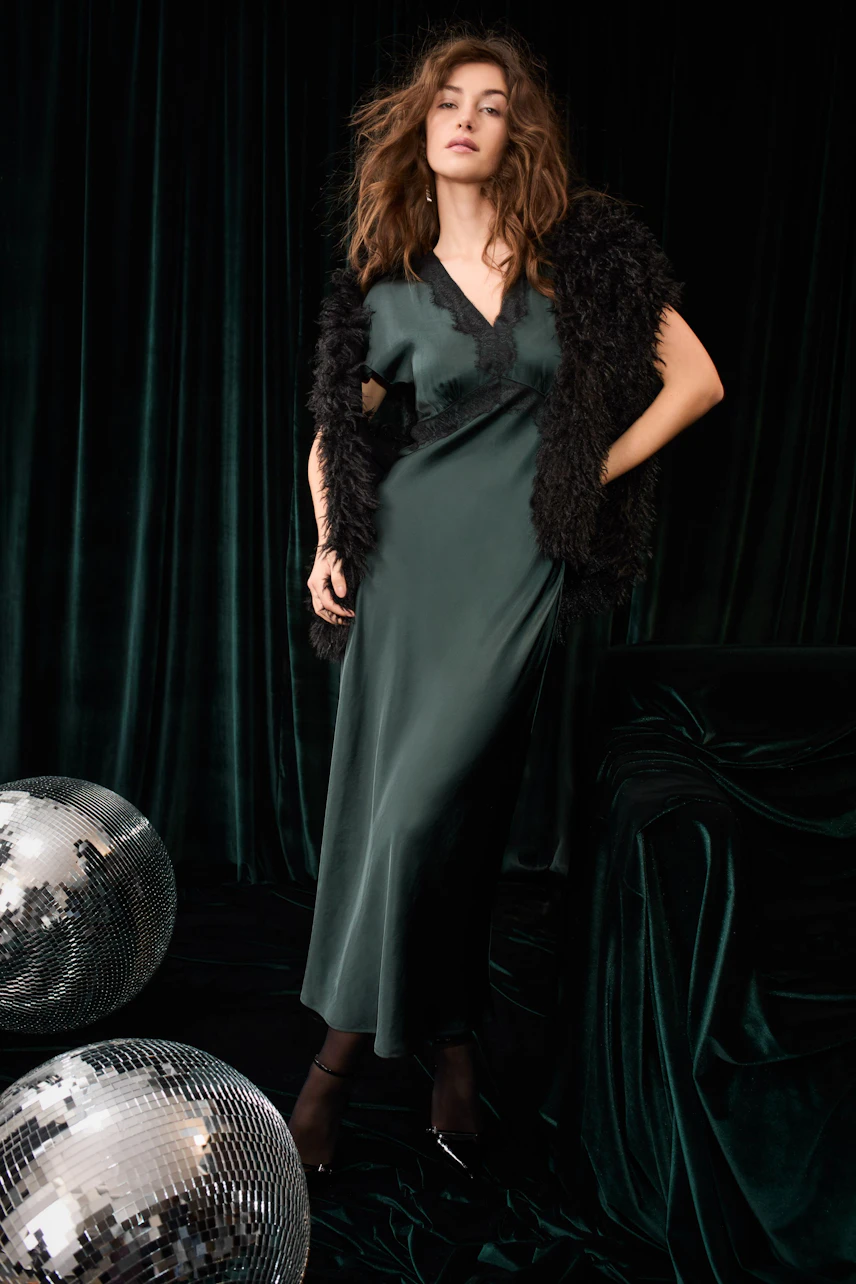 Woman in a sleek dress with a black fur shawl, standing confidently beside disco balls.