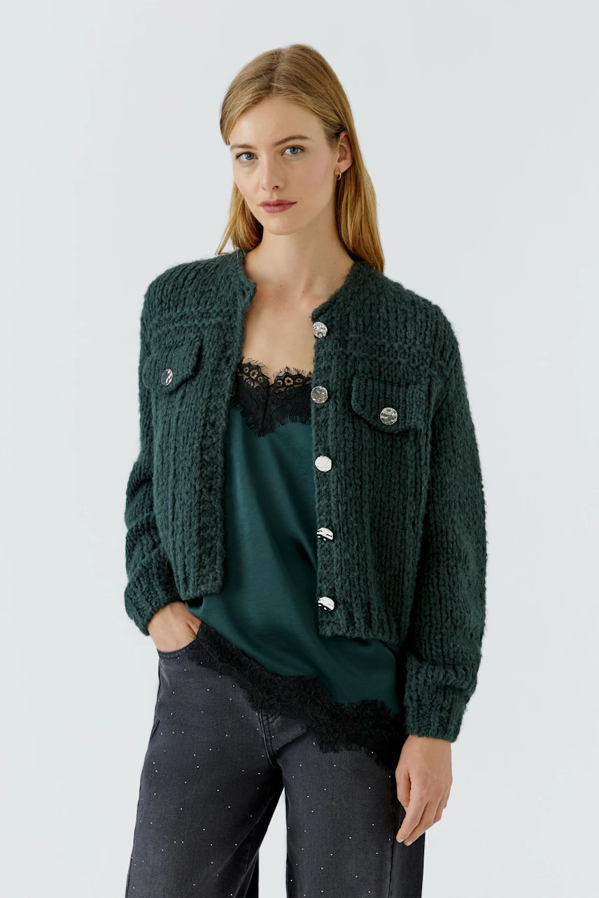 Strickjacke: Frau in grüner Strickjacke mit Knöpfen, kombiniert mit dunklem Top und Hose.