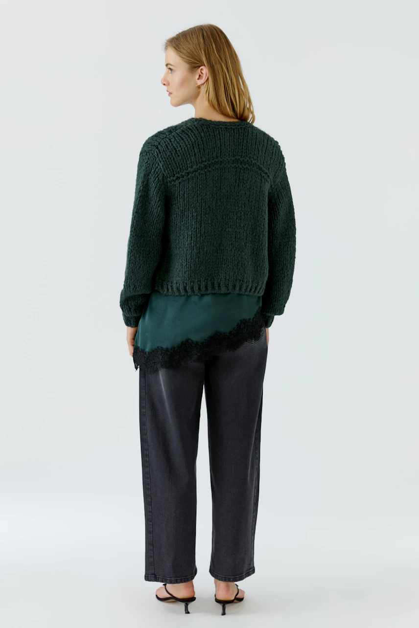 Frau in grüner Strickjacke, kombiniert mit schwarzer Hose und grüner Bluse, von hinten gesehen.