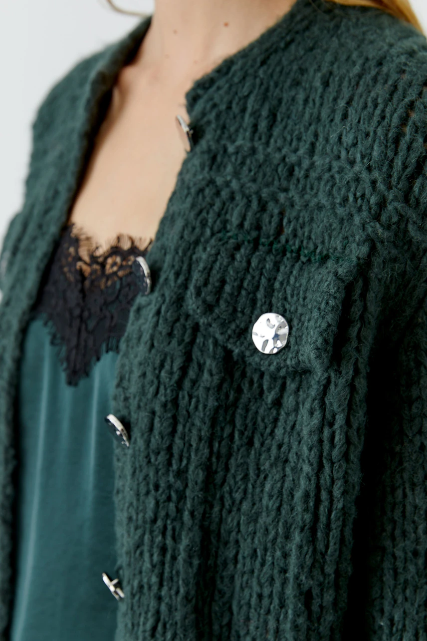 Frau in grüner Strickjacke mit Knöpfen, darunter ein Spitzen-Top. Die Jacke hat eine grobe Struktur.