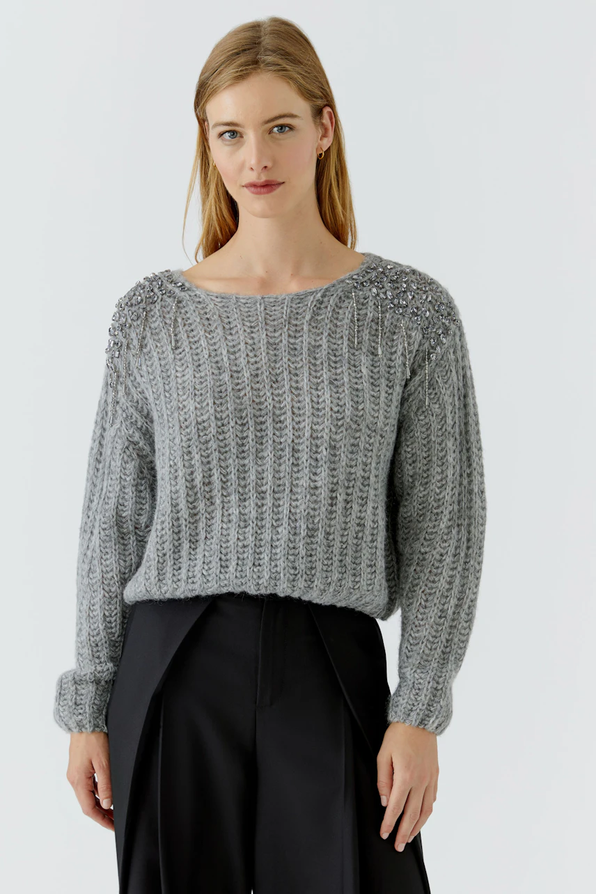 Grauer Damen-Pullover: Frau im grob gestrickten Pullover mit Ziersteinen, kombiniert mit schwarzer Hose.
