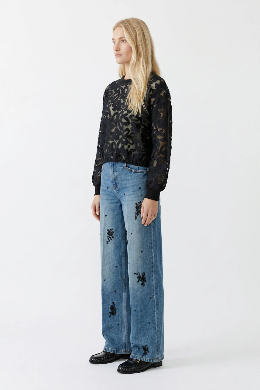 Damen-Pullover: Frau im schwarzen Spitzenpullover mit floralen Mustern und blauer Jeans steht seitlich.