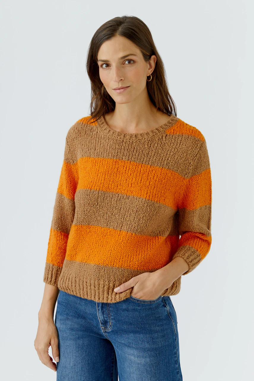 Frau im gestreiften Pullover in Orange und Braun, kombiniert mit blauen Jeans, schaut freundlich.
