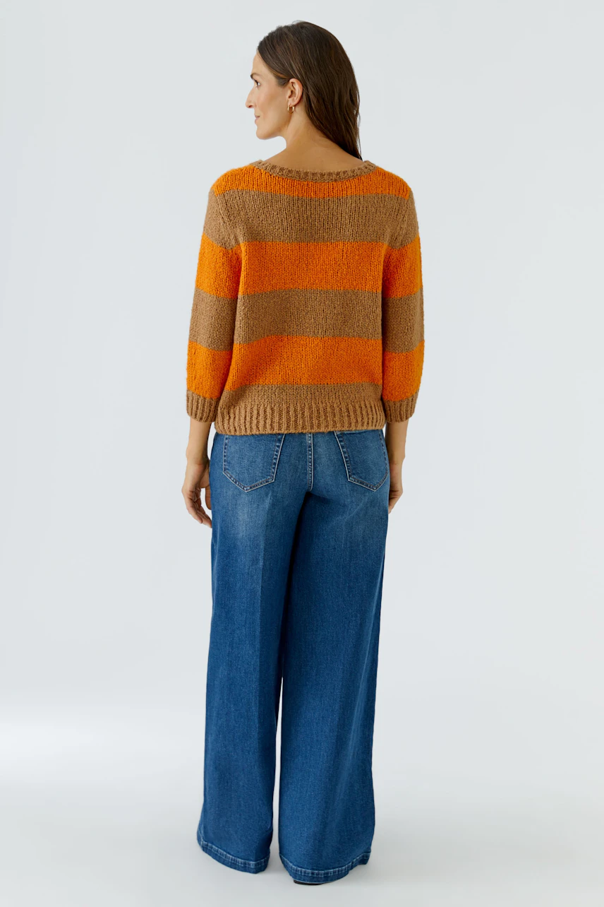 Frau im gestreiften Pullover in Orange und Braun, kombiniert mit blauer Jeans, von hinten gesehen.