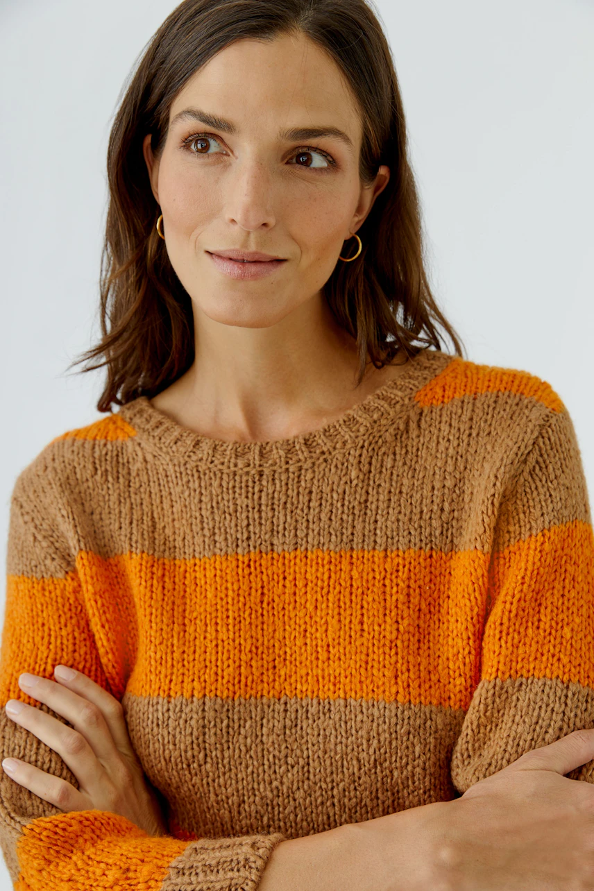 Frau im gestreiften Pullover in Orange und Braun, lässig und modern, mit verschränkten Armen.
