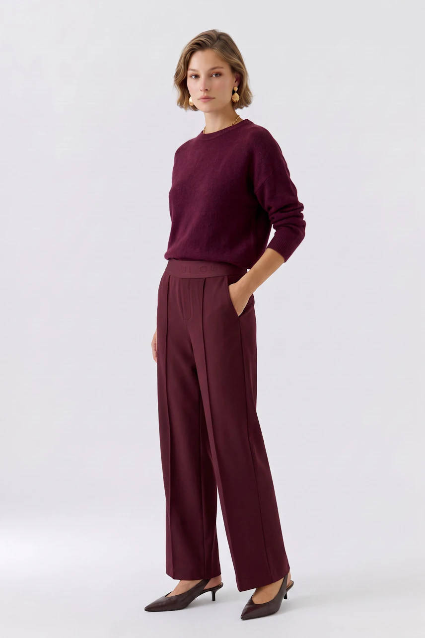 Woman in burgundy pants and matching pullover, standing confidently with hands in pockets.
