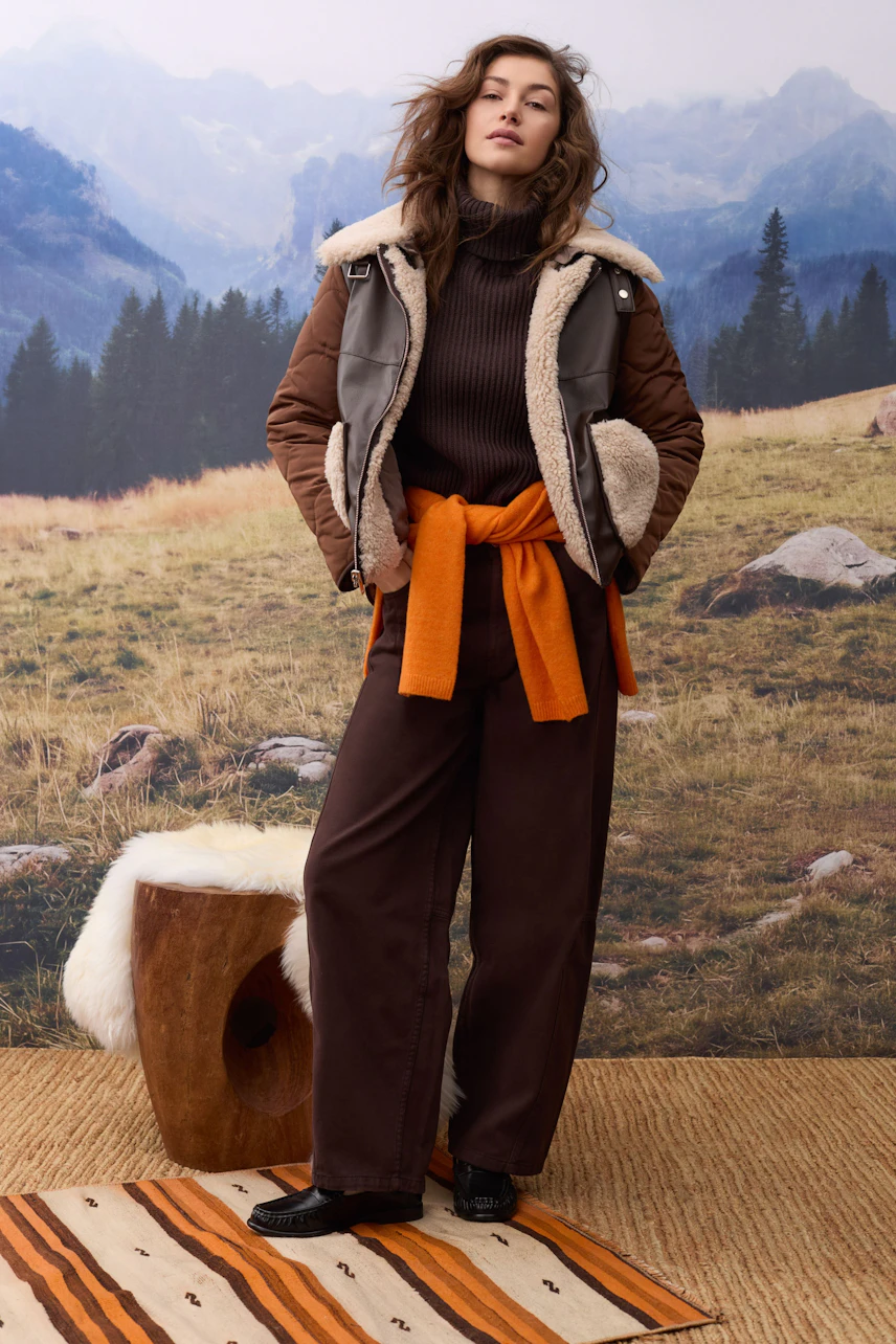 Frau im braunen Pullover mit orangefarbenem Schal, brauner Hose und Jacke vor Bergkulisse.