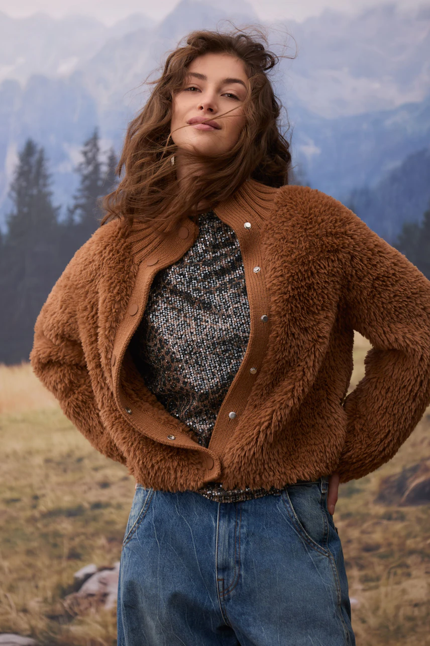 Frau in brauner Strickjacke: Lässige Strickjacke über gemustertem Oberteil und Jeans in natürlicher Umgebung.