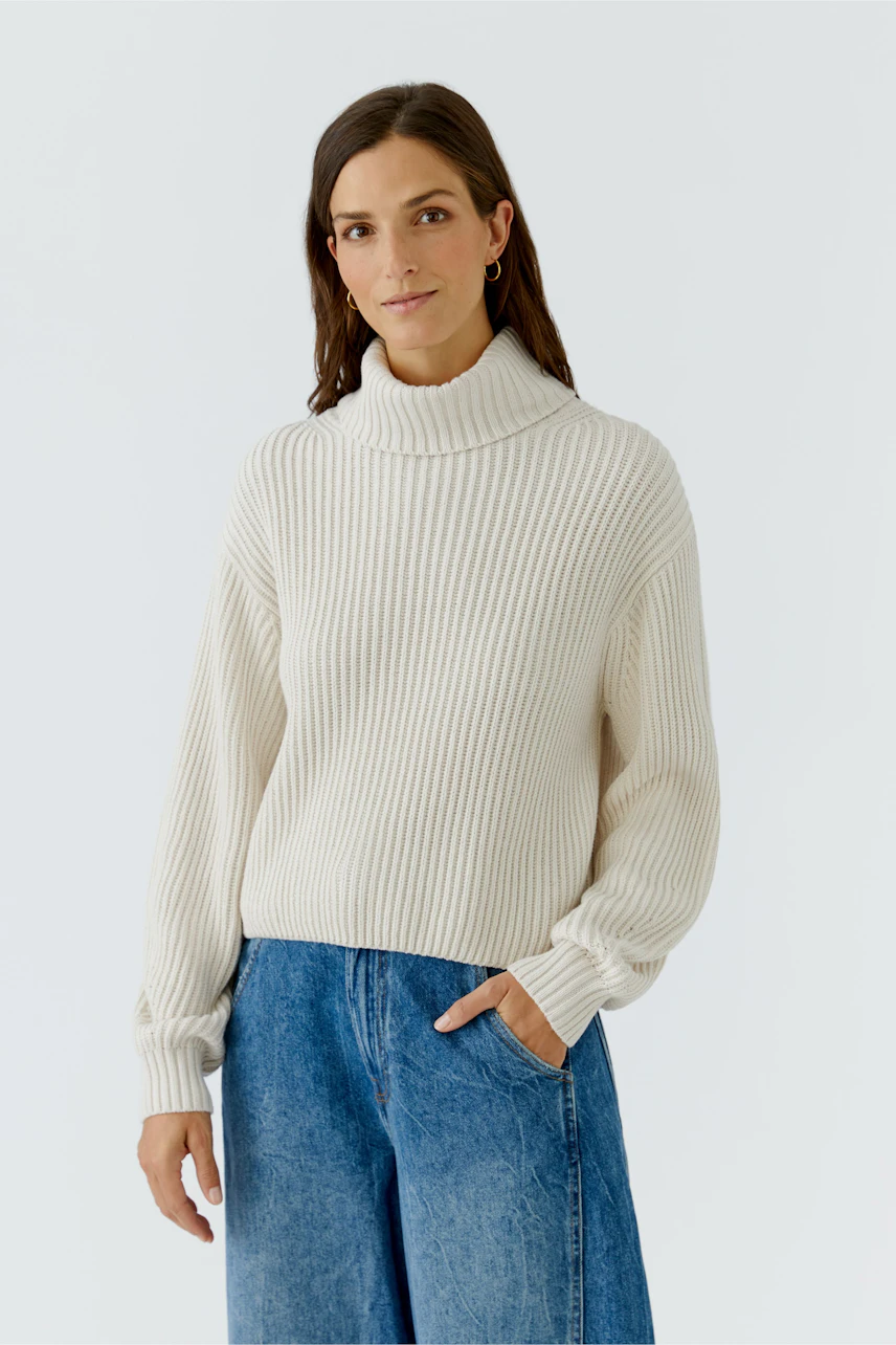 Frau im cremefarbenen Pullover mit Rollkragen, kombiniert mit blauer Jeans, lässig und modern.