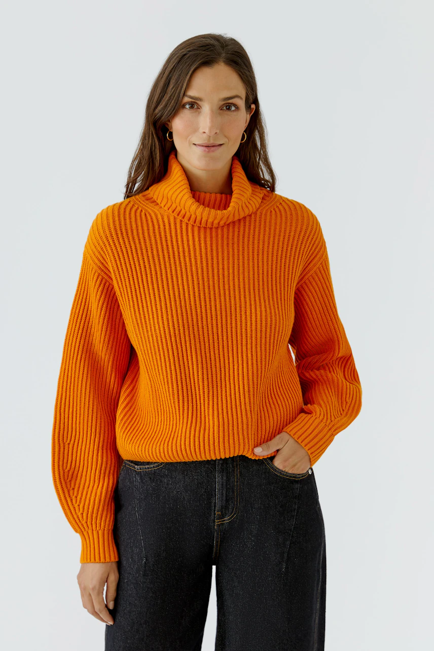 Frau im orangefarbenen Pullover mit Rollkragen, kombiniert mit dunkler Jeans, lächelt dezent.
