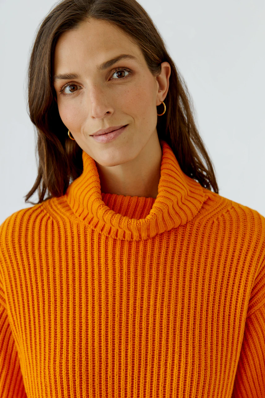 Frau im orangefarbenen Pullover mit Rollkragen, lächelt dezent. Ideal für einen warmen, modischen Look.