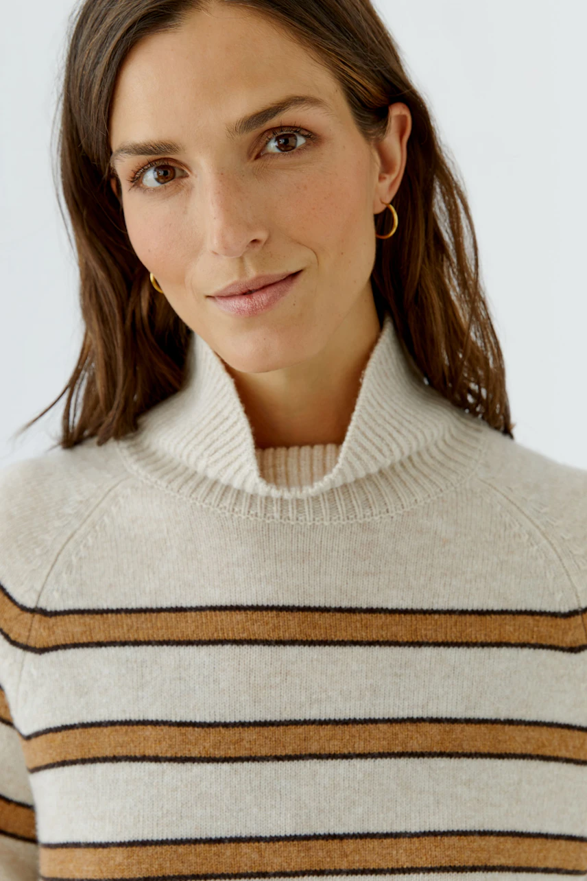 Frau im gestreiften Pullover mit Rollkragen, lächelt dezent. Der Pullover hat braune und beige Streifen.