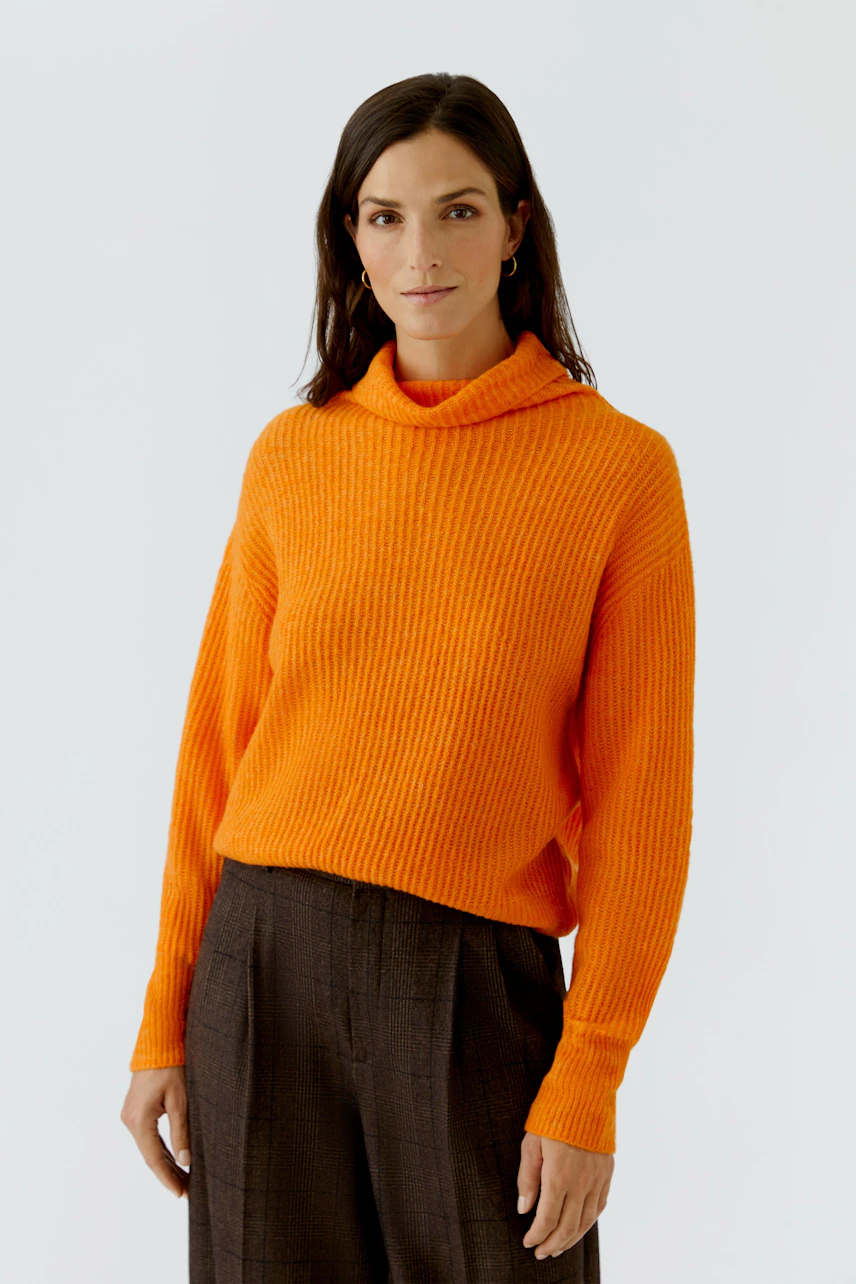 Frau im leuchtend orangefarbenen Pullover, kombiniert mit dunkler Hose, steht selbstbewusst.