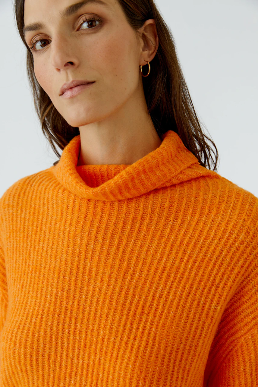 Frau im leuchtend orangefarbenen Pullover mit Rollkragen, schaut selbstbewusst in die Kamera.