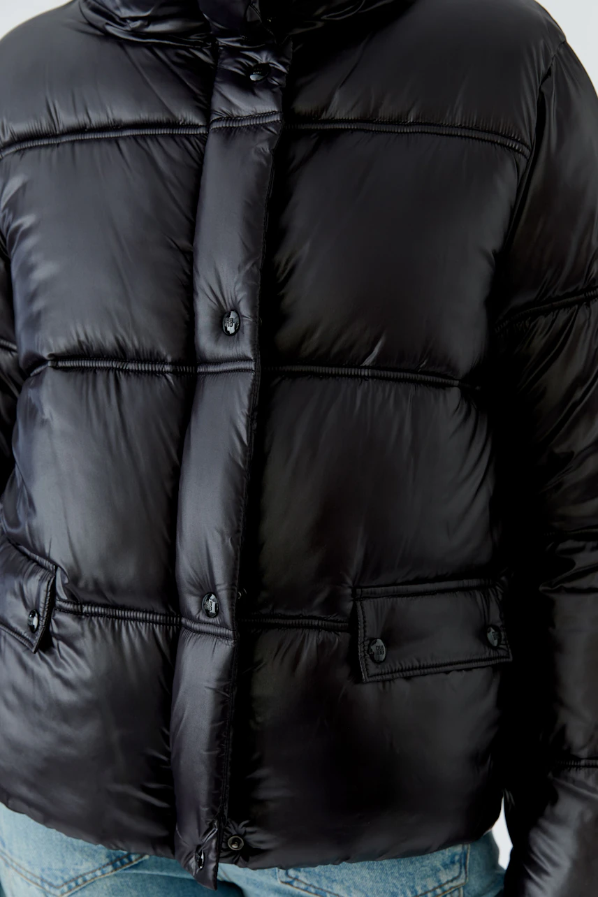 Schwarze Outdoorjacke: Frau in wattierter Jacke mit Kapuze und Knöpfen, kombiniert mit blauer Jeans.