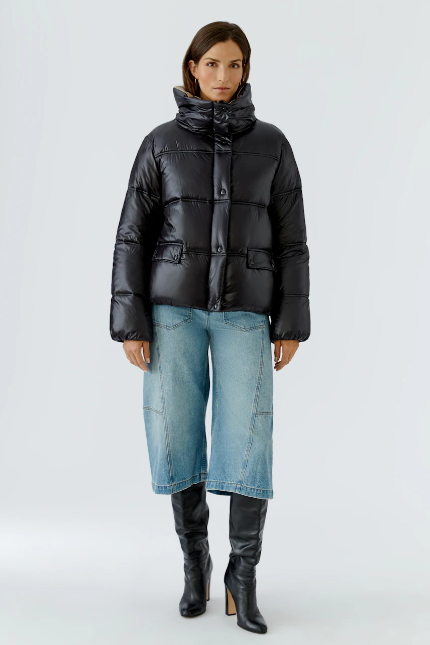 Damen-Outdoorjacke: Frau in schwarzer Steppjacke, blauer Jeans und schwarzen Stiefeln steht selbstbewusst.