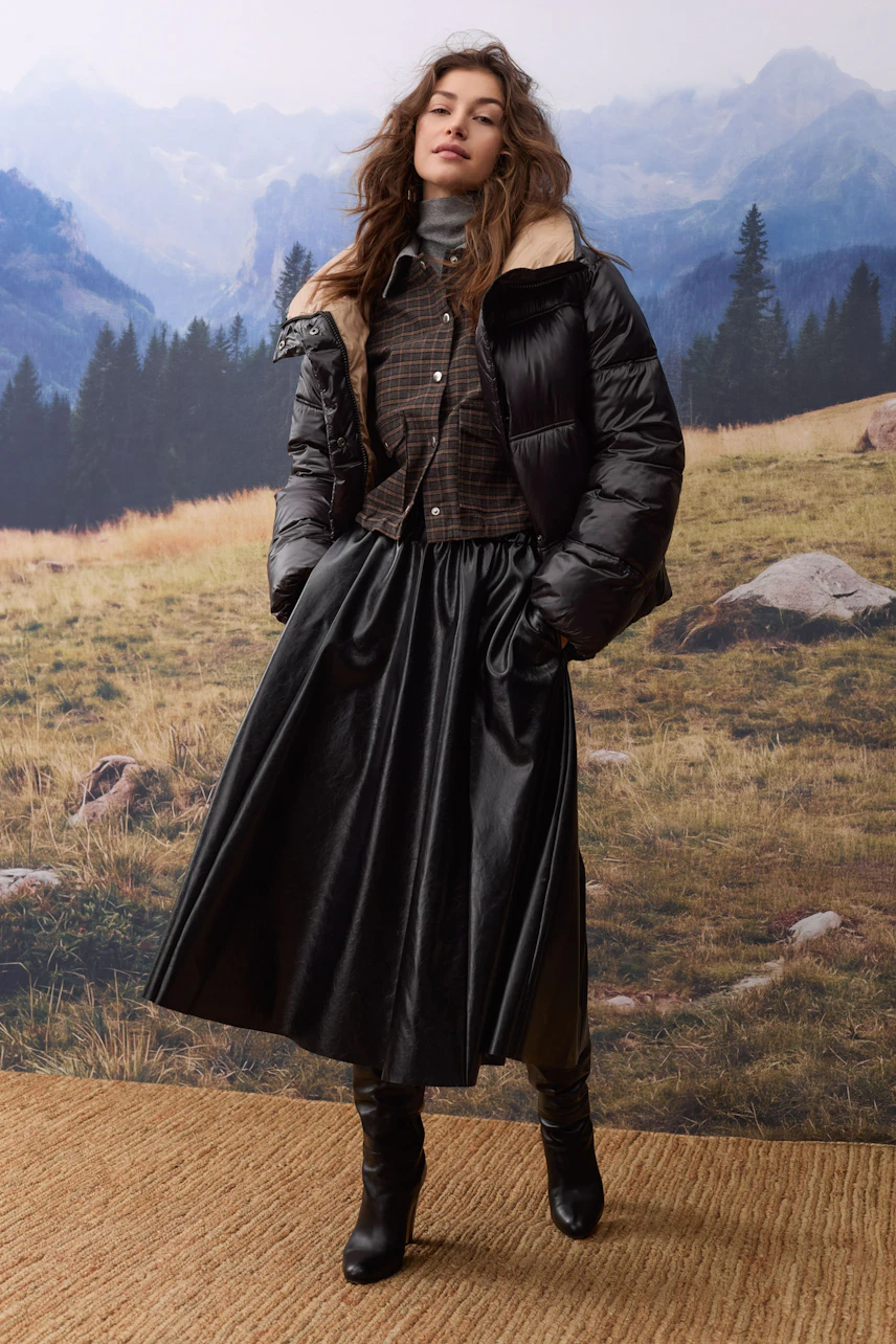 Outdoorjacke: Frau in schwarzer Steppjacke mit Fellkragen, kombiniert mit kariertem Hemd und langem Rock.