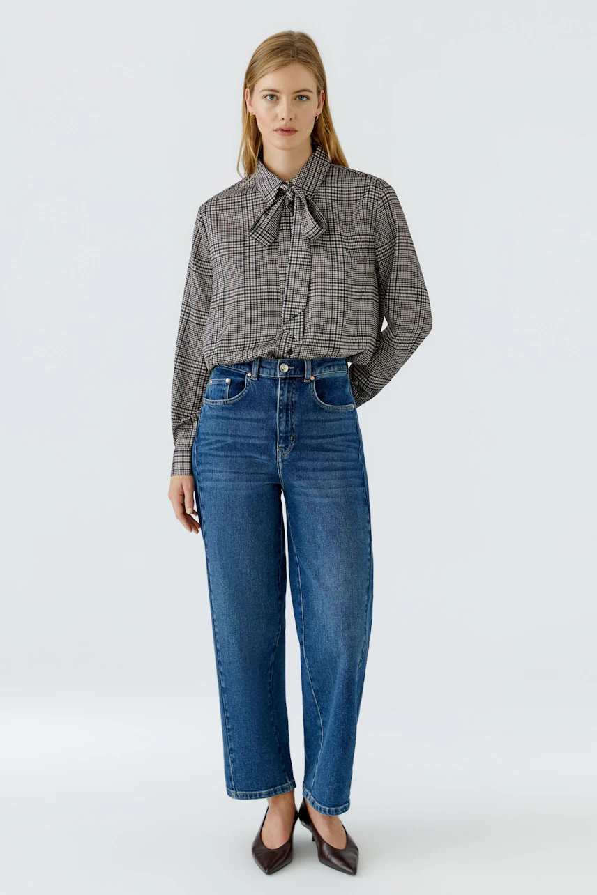 Woman in checkered pussy-bow blouse and blue jeans, standing confidently.
