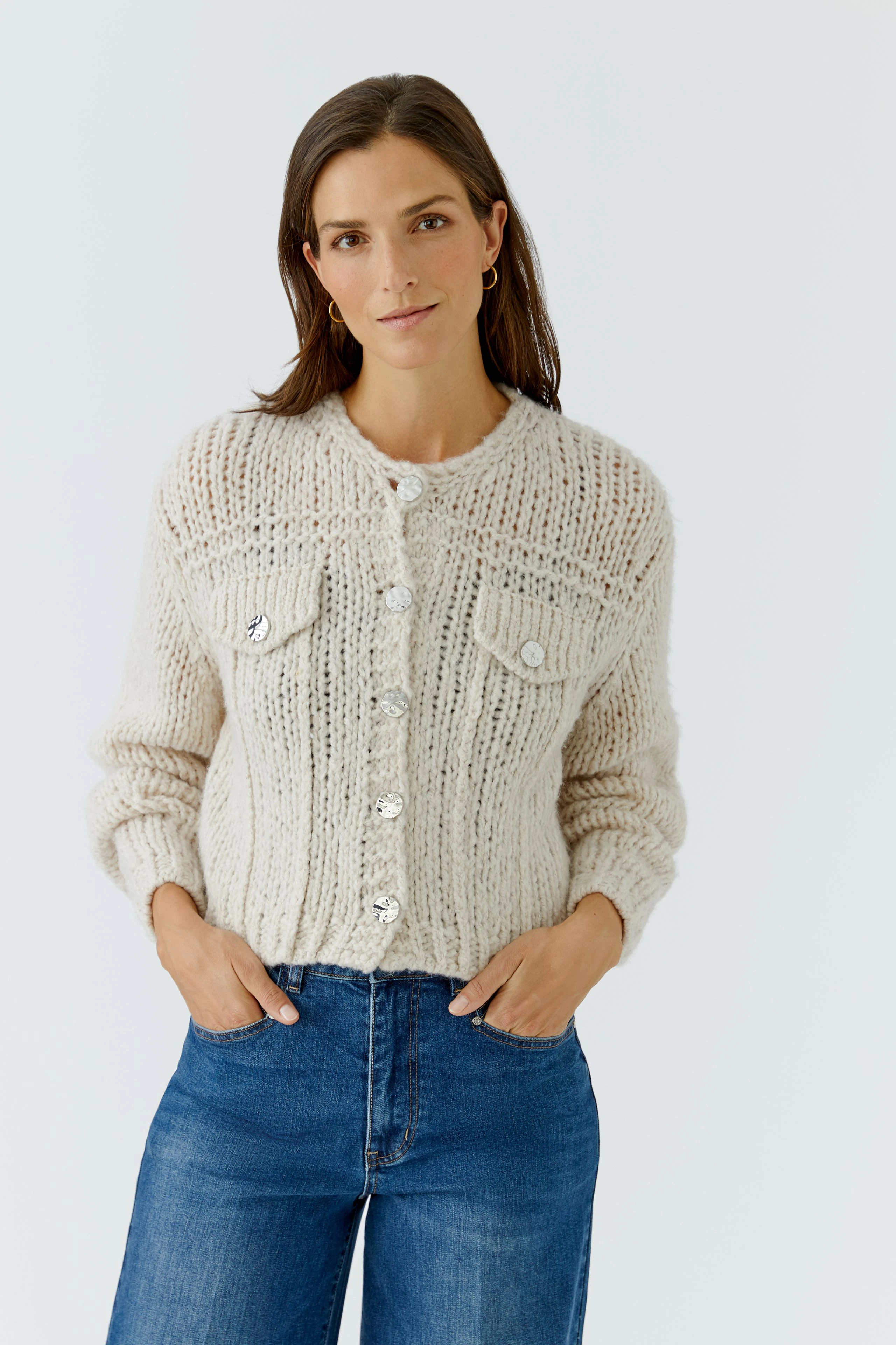 Woman in cozy knit cardigan with button details, hands in pockets, wearing blue jeans, looking relaxed.