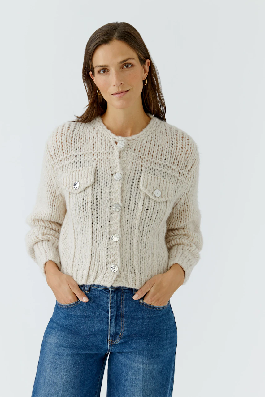 Woman in cozy knit cardigan with button details, hands in pockets, wearing blue jeans, looking relaxed.
