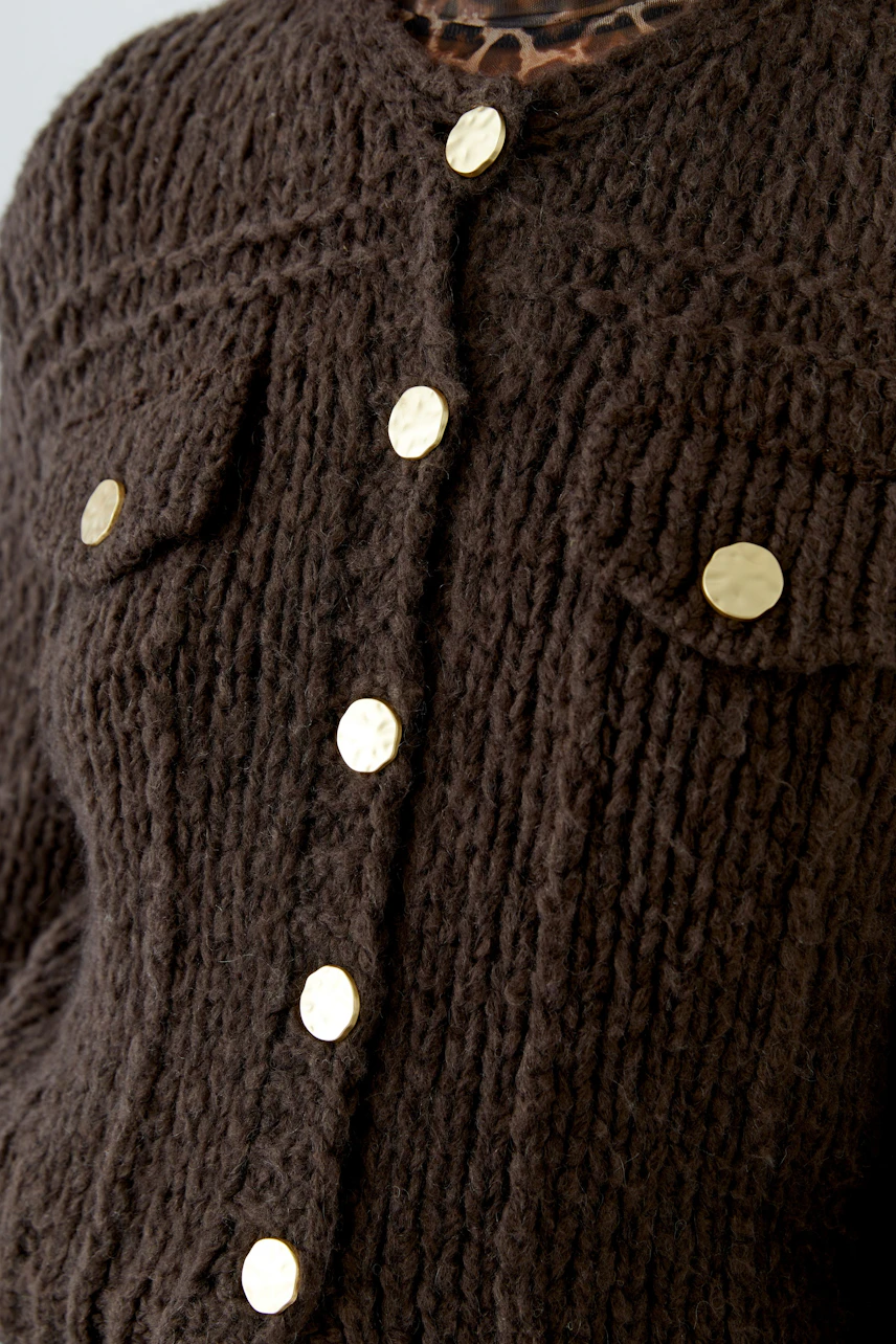 Chunky knit cardigan with gold buttons, close-up of textured fabric. No model visible.