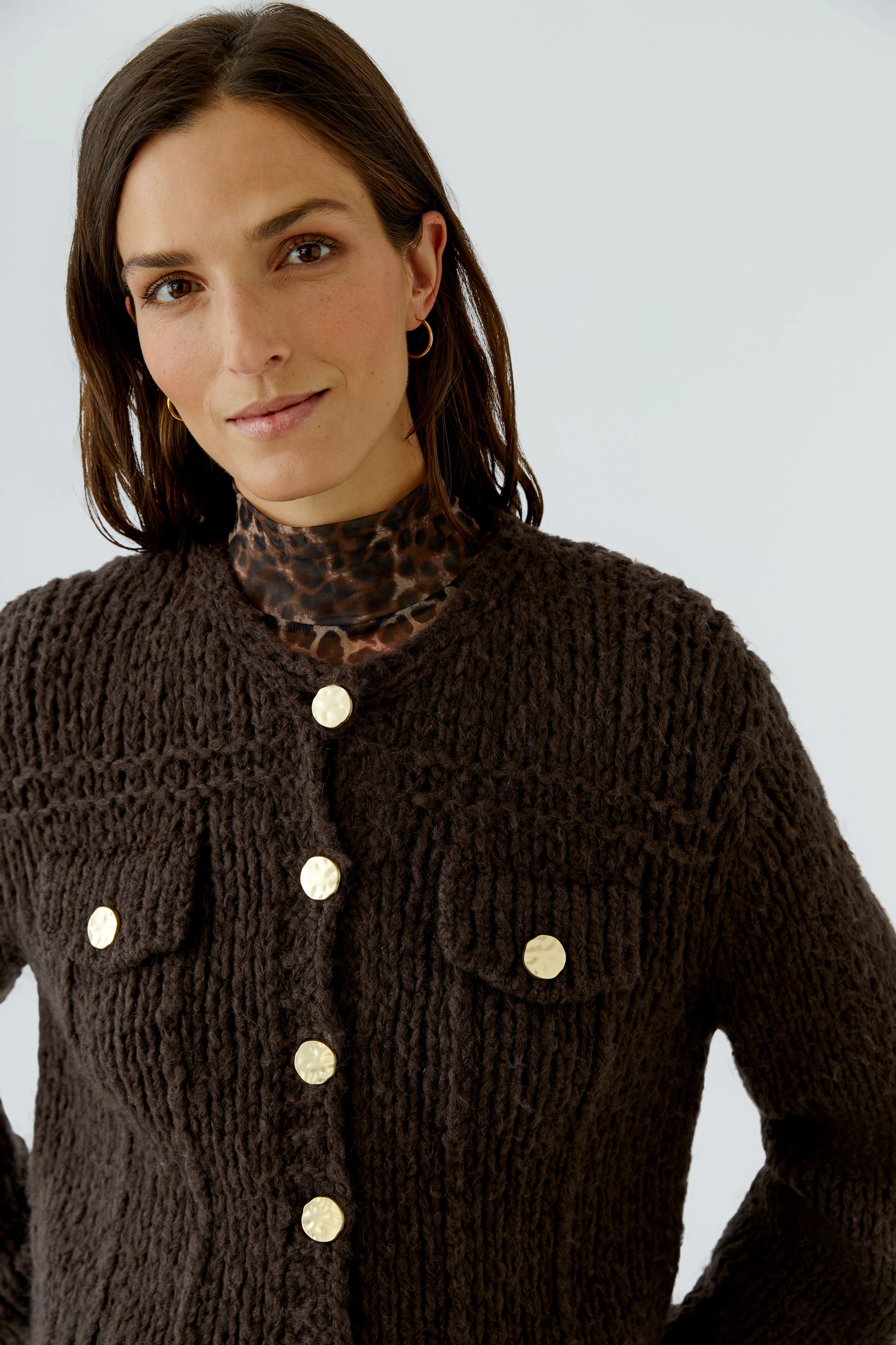 Woman in a chunky knit cardigan with gold buttons, layered over a leopard print top, looking confidently.