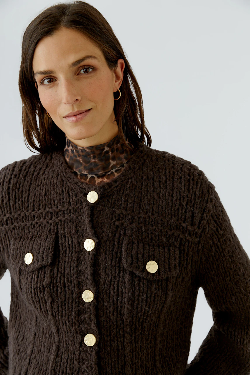 Woman in a chunky knit cardigan with gold buttons, layered over a leopard print top, looking confidently.