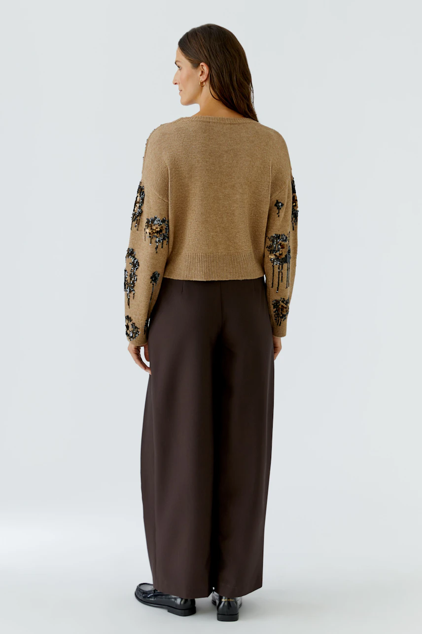 Damen-Pullover: Frau im beigen Pullover mit floralen Details, kombiniert mit brauner Hose, von hinten gezeigt.