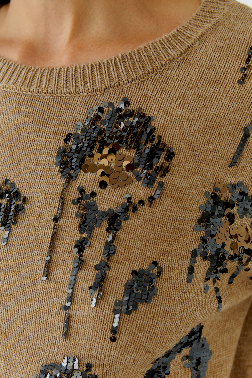 Damen-Pullover: Frau trägt beige Pullover mit schwarzen Paillettenmustern, Nahaufnahme des Designs.