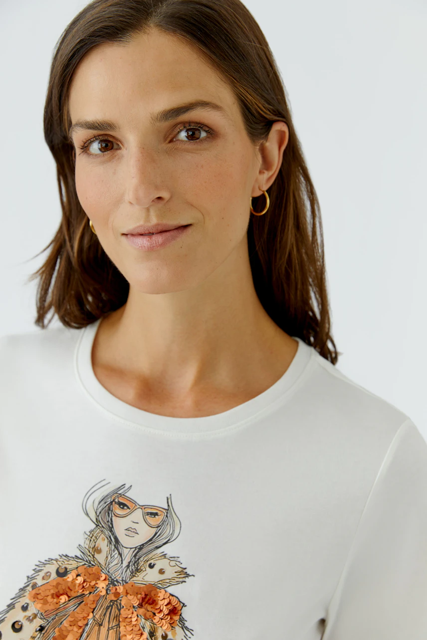 Casual women's T-shirt: Woman in white T-shirt with artistic print and gold hoop earrings, smiling softly.