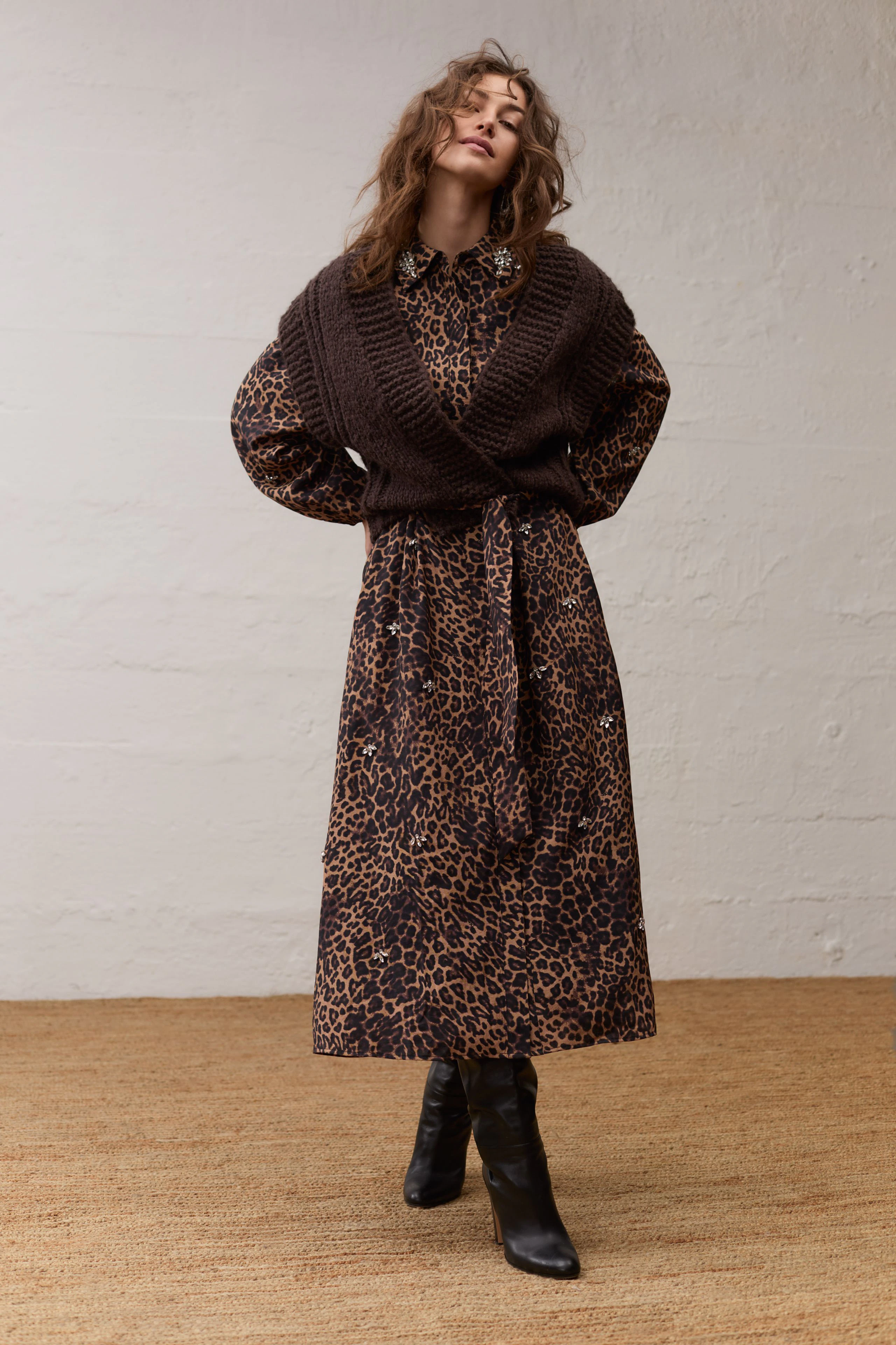 Woman in leopard print maxi dress with brown knit vest, standing confidently in black boots.