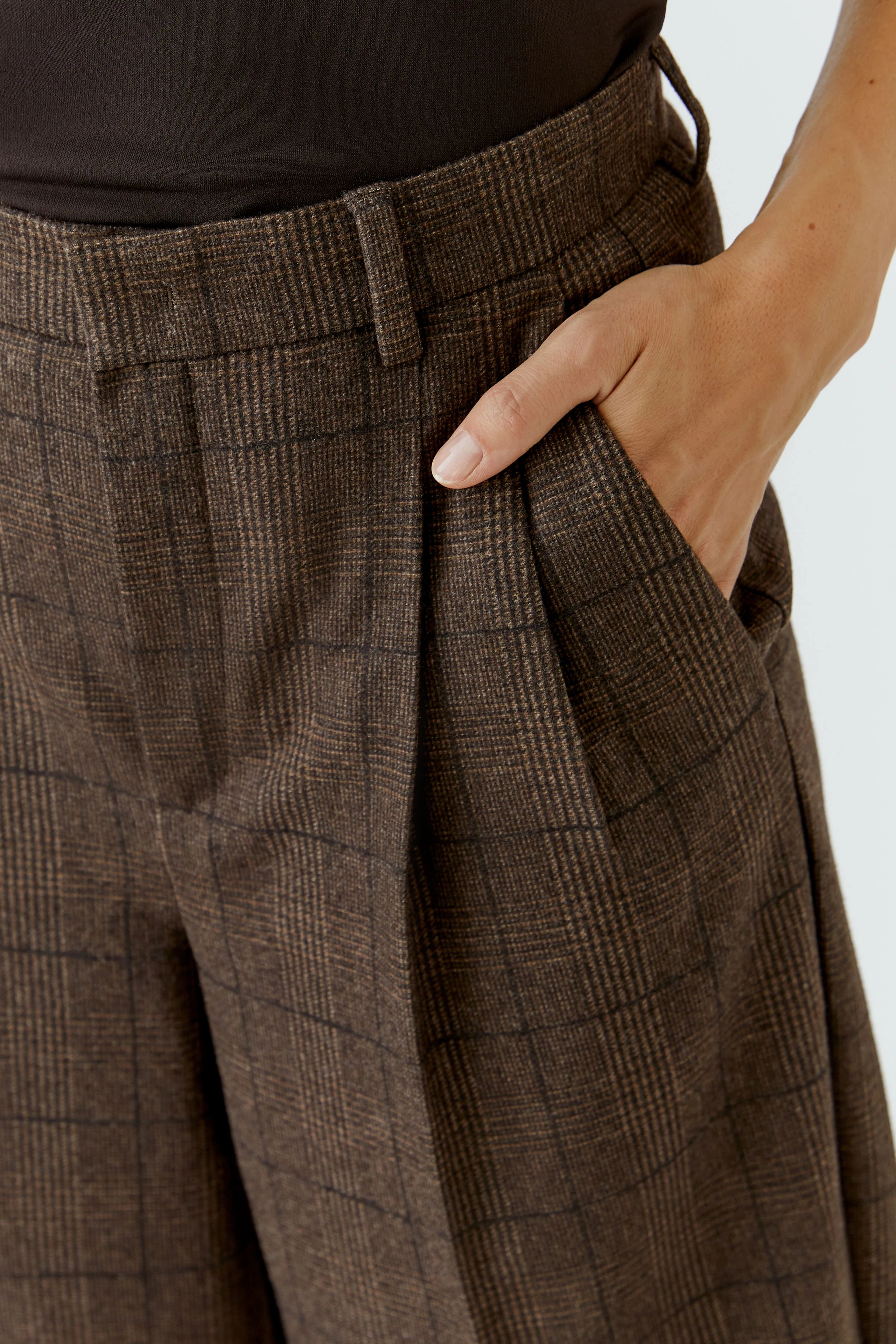 Woman in brown pleated trousers with a check pattern, hand in pocket, wearing a black top.
