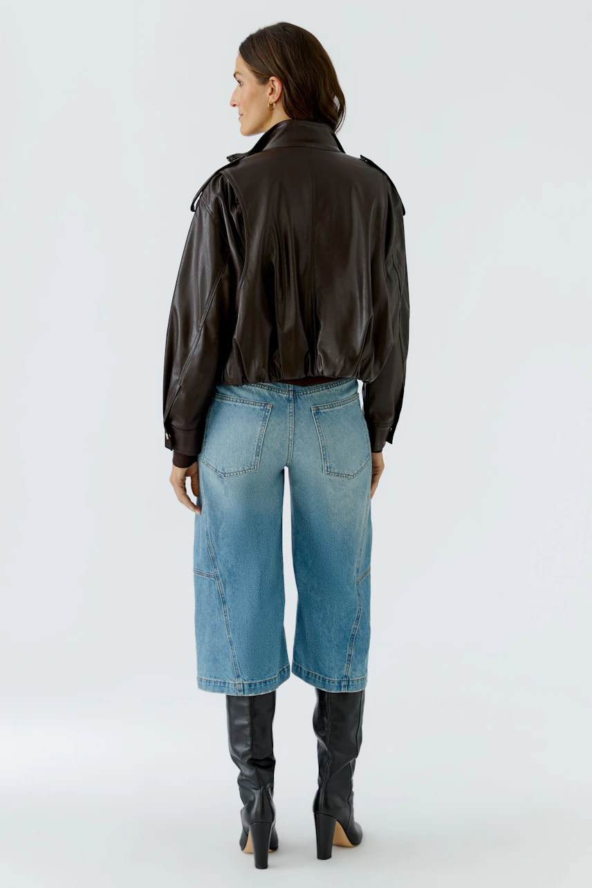 Lederimitatjacke: Frau in brauner Jacke, blauer Jeans und schwarzen Stiefeln, von hinten gesehen.
