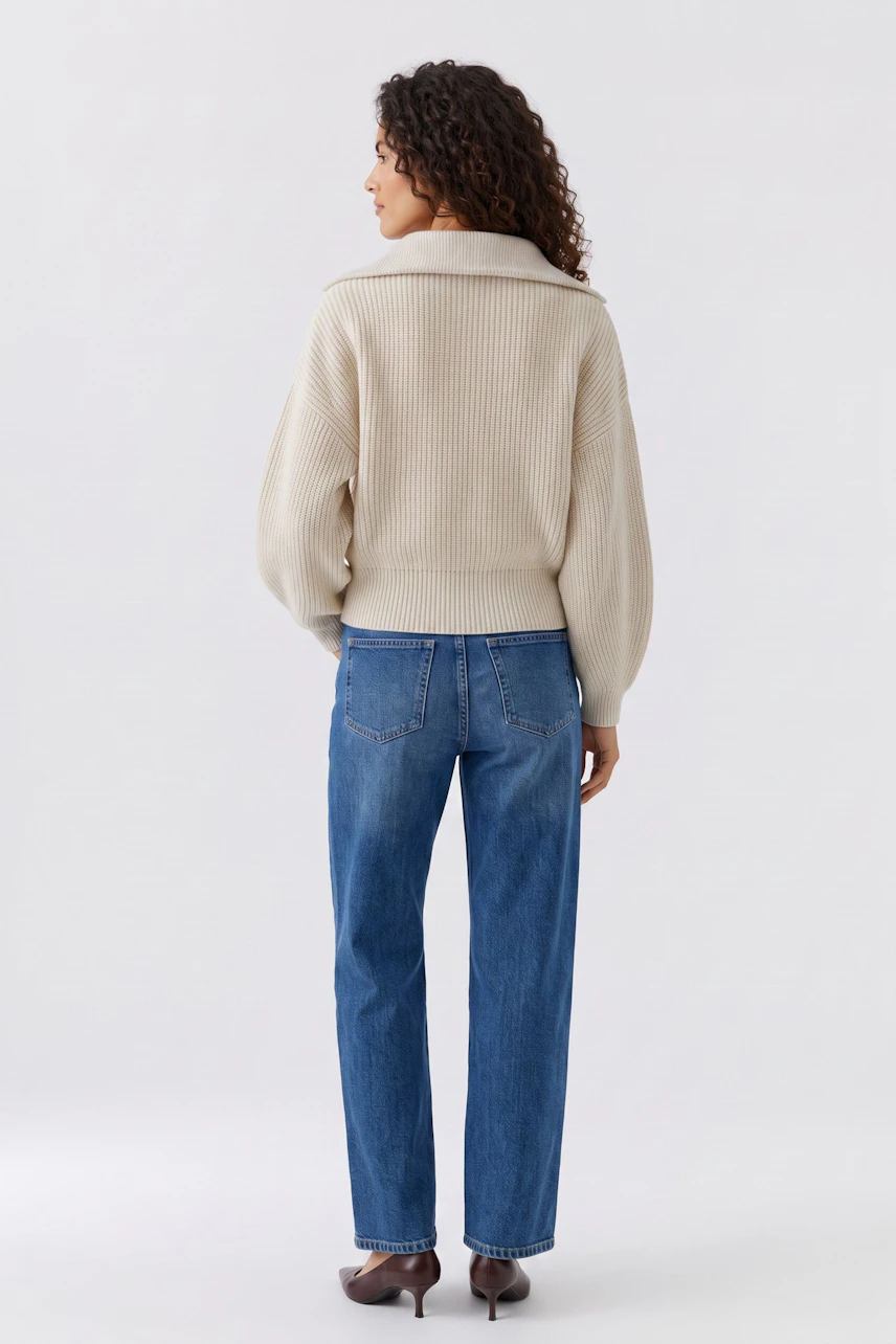 Damen-Pullover: Frau im cremefarbenen Pullover und blauer Jeans, von hinten gesehen, mit lockigem Haar.
