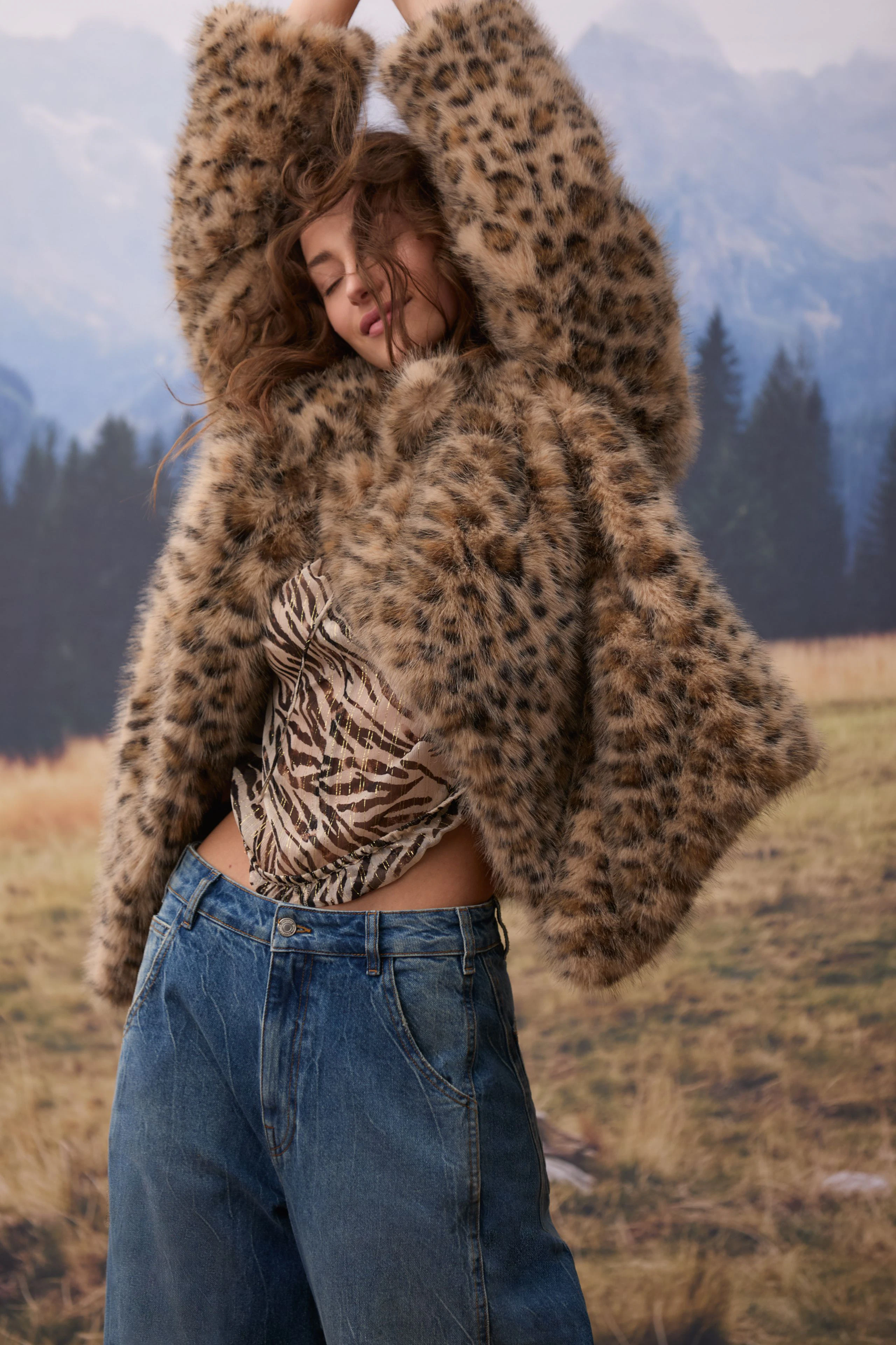 Woman in a leopard print blouse and jeans, arms raised, in a natural outdoor setting.