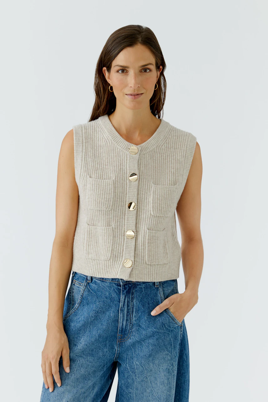 Strickweste: Frau in beiger Strickweste mit goldenen Knöpfen und blauer Jeans, lässig und modern.